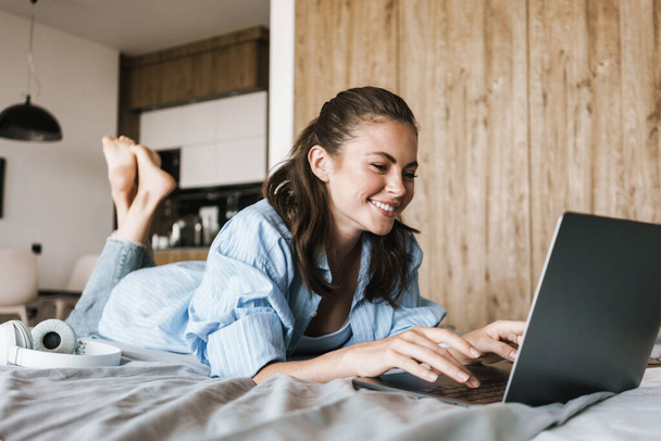 Image of a young happy smiling girl on a bed in bedroom indoors at home using laptop computer - Valokuva, kuva