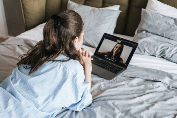 Photo of focused nice girl making video call on laptop while lying on bed in living room - Fotó, kép