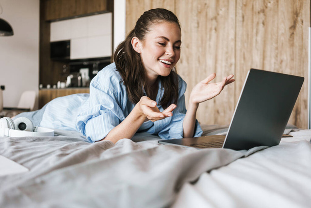 Photo of happy nice girl smiling and making video call on laptop while lying on bed in living room - Valokuva, kuva