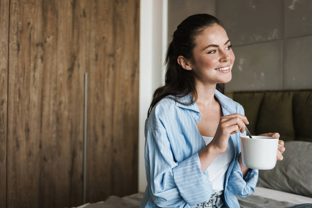 Image of a young cheerful happy pleased girl indoors at home drinking coffee - Foto, Imagen
