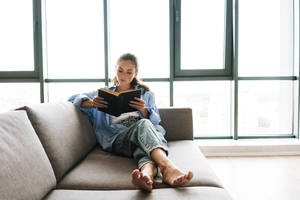 Image of a young concentrated girl indoors at home sitting on sofa while reading book - Foto, Bild