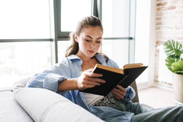 Image of a young concentrated girl indoors at home sitting on sofa while reading book - Foto, imagen