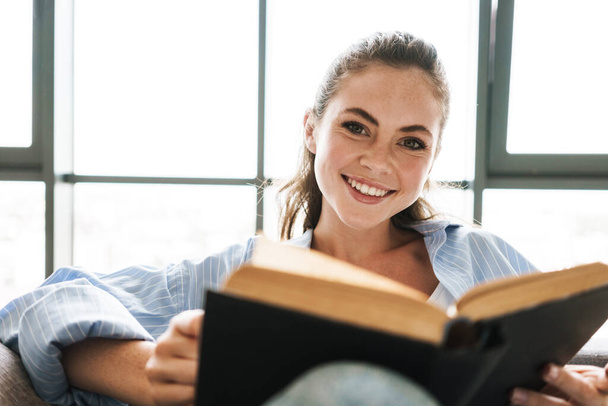 Image of a young happy positive girl indoors at home sitting on sofa while reading book - Фото, зображення