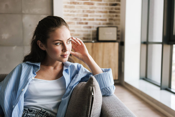 Photo of pleased nice girl looking aside while sitting on couch in living room - Foto, Imagem