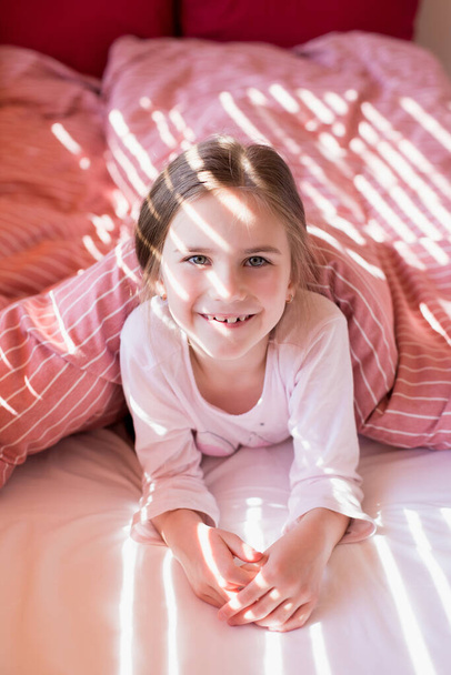 Yatakta tatlı küçük bir kız sabah uyanır ve neşeyle gülümser. Güneşin ışınları jaluzilerden çocuğun yüzüne düşer. Karantina sırasında evde vakit geçirmek. Ev rahatlığı. Tasasız çocukluk - Fotoğraf, Görsel