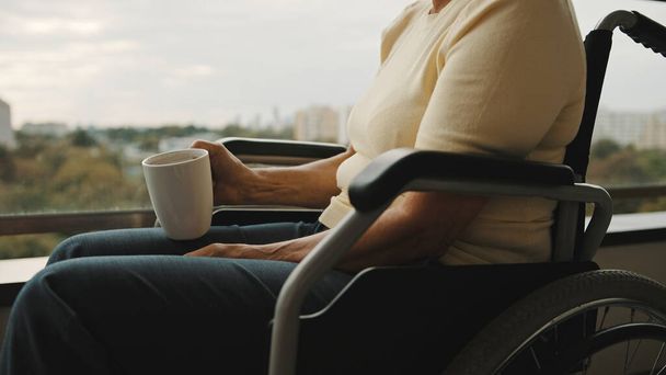 ηλικιωμένη γυναίκα σε αναπηρική καρέκλα στη βεράντα κρατώντας ένα φλιτζάνι τσάι ή καφέ. Χωρίς πρόσωπο - Φωτογραφία, εικόνα