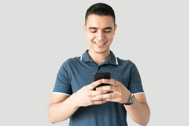Jovem bonito usando seu smartphone para verificar nas mídias sociais em um estúdio e sorrindo - Foto, Imagem
