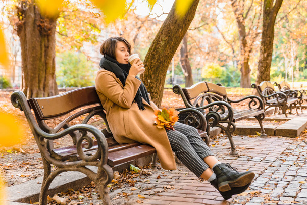 žena sedí na lavičce v podzimním městském parku a pije kávu podzimní sezóny - Fotografie, Obrázek