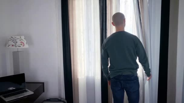 young man stands in front of the window, opens the curtains, looks at the street, the concept of business travel, tourism, loneliness, stay at home, quarantine - Πλάνα, βίντεο