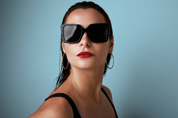 Colourful studio portrait of a young woman wearing dark sunglasses, posing over blue background. - Photo, Image