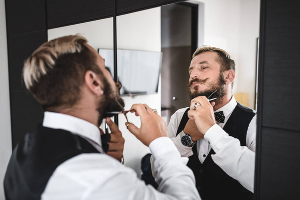 Young man trims his beard with scissors while standing in front of a mirror - Photo, Image