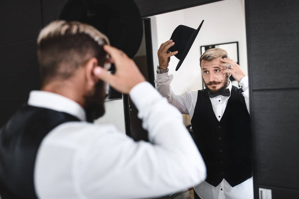 Stylish man with mustache and beard puts on hat in front of the mirror - Photo, Image