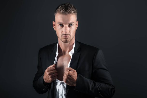 Confident elegant handsome young man unbuttons his shirt and stands on a dark background in the studio in a beautiful suit - Fotografie, Obrázek