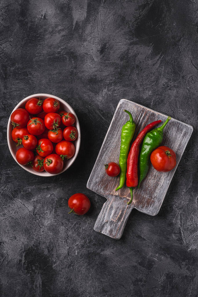 Fresh ripe tomatoes in bowl near to old wooden cutting board with hot chili peppers, dark stone concrete background, top view - Photo, Image