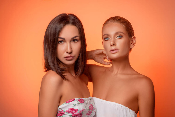 portrait of two attractive caucasian women blond and brunette isolated on orange studio shot and shoulders looking at camera - Fotó, kép