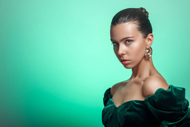 Portrait of a Caucasian girl in modern design hoop earrings and green off-shoulder dress. Studio background - Foto, imagen