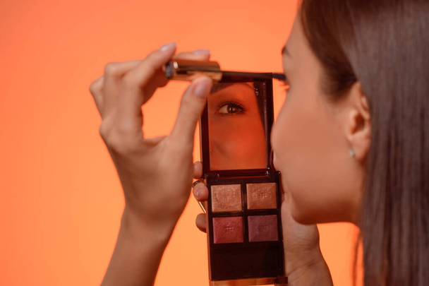 An elegant brunette holds a makeup mirror in her hands and dresses up, paints her eyelashes with mascara. Studio Orange Background. Copy Space - 写真・画像