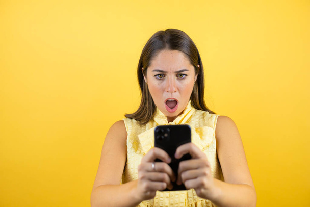 Junge schöne Frau auf isoliertem gelben Hintergrund plaudert mit ihrem Handy und überrascht - Foto, Bild