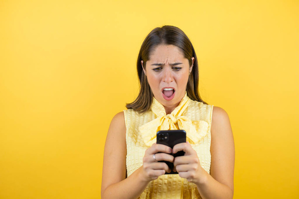 Mladá krásná žena přes izolované žluté pozadí chatování se svým telefonem a překvapený - Fotografie, Obrázek