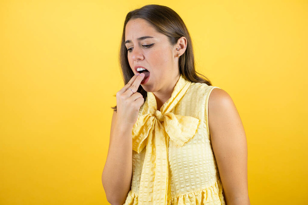 Junge schöne Frau auf isoliertem gelben Hintergrund angewidert mit der Hand im Mund - Foto, Bild