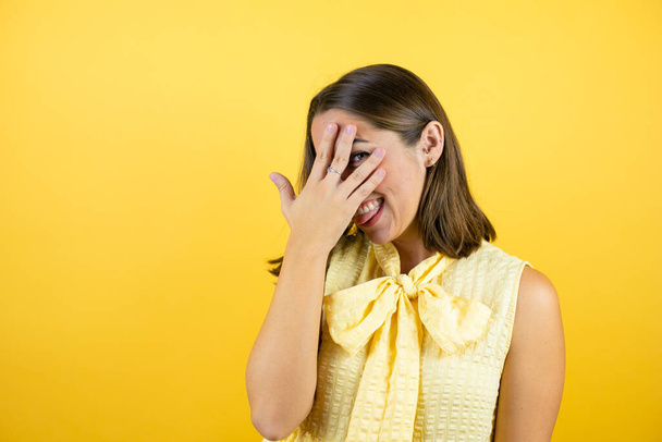 Young beautiful woman over isolated yellow background peeking in shock covering face and eyes with hand, looking through fingers with embarrassed expression - Photo, Image