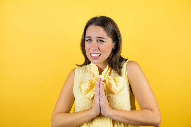 Молодая красивая женщина на изолированном желтом фоне умоляя и молясь руками вместе с выражением надежды на лице очень эмоциональным и обеспокоенным - Фото, изображение