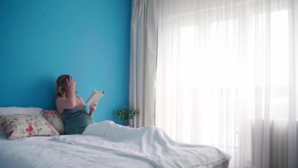 Těhotná žena v polosedící poloze ve své posteli a vypadá šťastně, když čte knihu. Má jednu ruku na břiše.. - Záběry, video