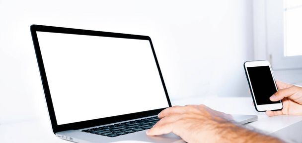 Persona que trabaja desde su casa en su escritorio usando una computadora portátil y un teléfono móvil en un escritorio blanco. - Foto, imagen
