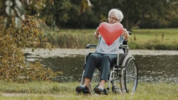 Nehrin kenarındaki tekerlekli sandalyede oturan yaşlı kadın elinde kalp şeklinde balon tutuyor. - Video, Çekim