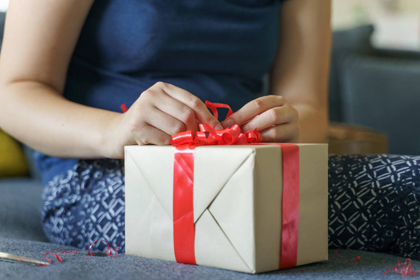 Close-up van onbekende blanke vrouw middendoorsnede en handen zitten op de bank thuis met het maken of openen van verpakt doos met rode stropdas lint - verjaardag of vakantie cadeau geven en ontvangen concept - Foto, afbeelding