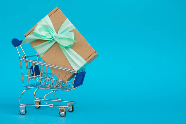 Shopping cart with huge gift in craft paper with a mint ribbon on blue background. Copy space - Photo, Image