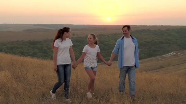 healthy smiling family holding hands walking in field at sunset in the mountains, daughter holding mom and dad by hands. happy child and parents walk in rays of beautiful sun, travel on vacation - Footage, Video