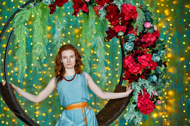 A young red haired woman in a turquoise dress stands at a cradle with red flowers against the background of a bokeh of yellow garland lights. - Photo, Image