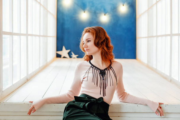 Egy vonzó vörös hajú fiatal nő pózol egy tágas szobában, közel egy fehér ablakhoz és egy kék falhoz, koszorúval.. - Fotó, kép