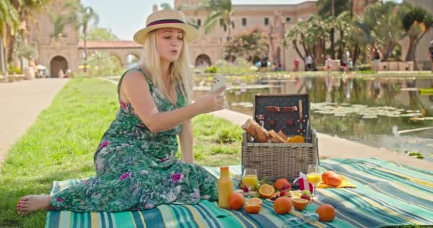 Aantrekkelijke blonde vrouw met behulp van mobiele telefoon tijdens de picknick bij de vijver in park 4K  - Video