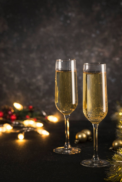 New year 2021 - two glasses of champagne on a dark background with bokeh from garlands. Classic alcoholic drink for a new year's holiday atmosphere. Vertical orientation with a copyspace. - Photo, image