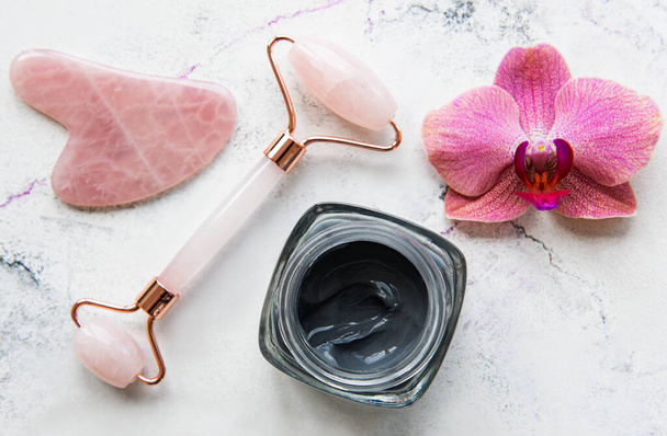 Skincare reggeli szépség rutin otthon fürdőszoba - arc jáde görgő masszírozó wellness kozmetikai terápia, maszk és orchidea virágok márvány háttér - Fotó, kép