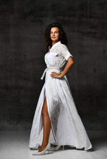 Elegant powerful brunette woman in white long lace dress with short sleeves and high heeled shoes stands looking at camera - 写真・画像
