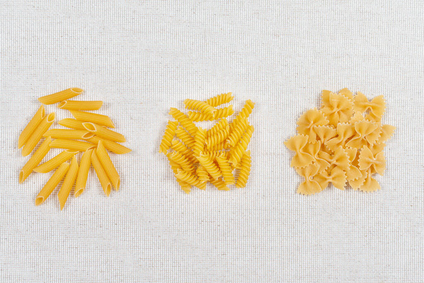Various raw pasta types on rustic linen fabric, top view. Various forms of pasta fusilli, penne and farfalle - Photo, Image