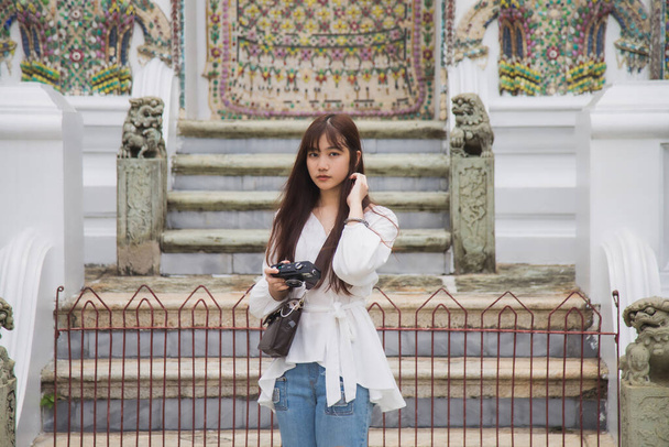 De cerca retrato joven mujer asiática en una camisa blanca sosteniendo una cámara retro y mirando a la cámara mientras viaja dentro de Wat Phra Kaew en un día de mal humor - Foto, Imagen