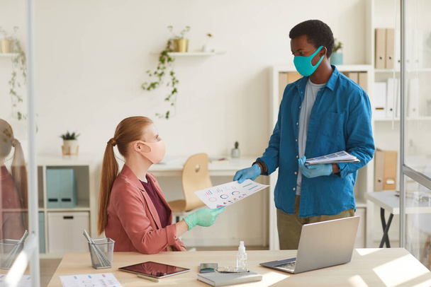 Portrait d'une jeune femme d'affaires portant un masque et des gants remettant des documents à un collègue afro-américain pendant qu'elle travaillait dans un bureau post-pandémique, photocopier - Photo, image