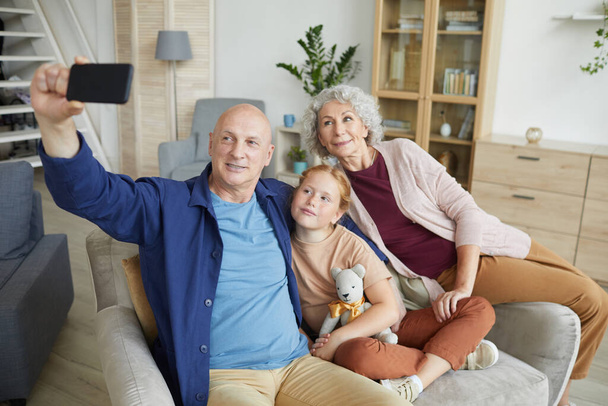 Retrato de pareja de ancianos modernos tomando foto selfie a través de teléfono inteligente con linda chica de pelo rojo en el interior del hogar, espacio de copia - Foto, imagen
