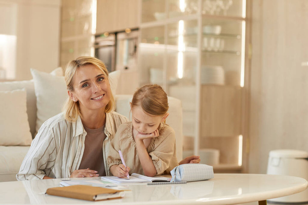 Warm-toned portrait of happy young mother smiling at camera while helping cute little girl drawing on studying at home, copy space - Photo, Image