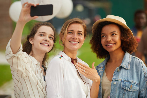 Waist up portrait of three beautiful young women taking selfie photo via smartphone during outdoor party in Summer - Photo, Image