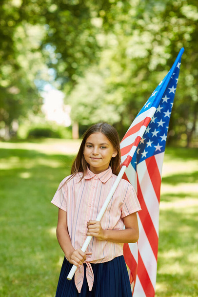 Vertical waist up portrait of smiling schoolgirl carrying American flag while standing outdoors in sunlight, copy space - Photo, Image