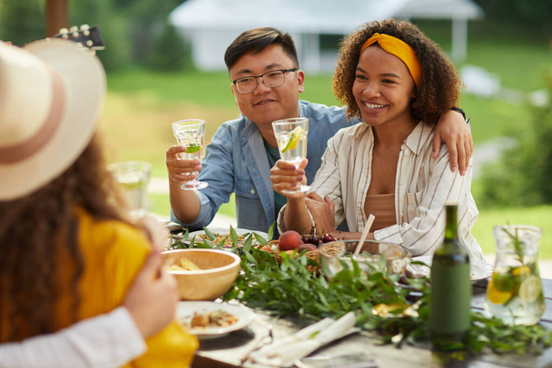 Retrato de mestiço jovem casal abraçando enquanto sentado mesa segurando bebidas e desfrutar de jantar com amigos ao ar livre na festa de verão, espaço de cópia - Foto, Imagem