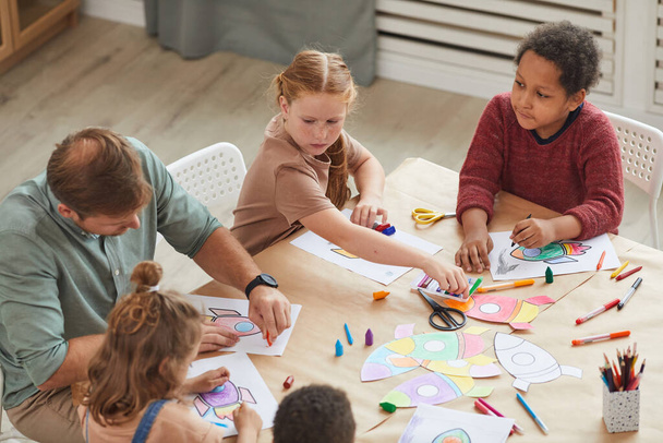 Vista de ángulo alto en un grupo multiétnico de niños que dibujan dibujos con lápices de colores mientras disfrutan de clases de arte y artesanía en preescolar o centro de desarrollo, espacio para copiar - Foto, Imagen