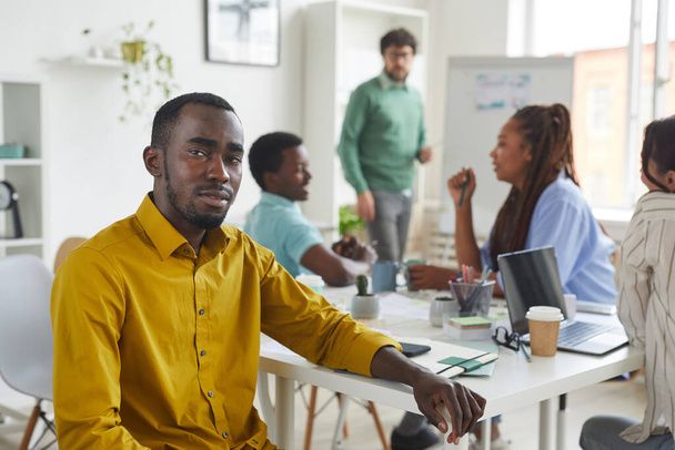 Portré kortárs afro-amerikai férfi nézi a kamerát, miközben ül az asztalnál, miközben találkozik az üzleti csapat a háttérben, másolja a helyet - Fotó, kép