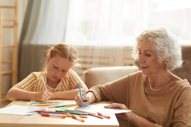 Warm toned portrait of smiling senior woman babysitting cute red haired girl and drawing together while sitting by coffee table in cozy living room - Photo, Image