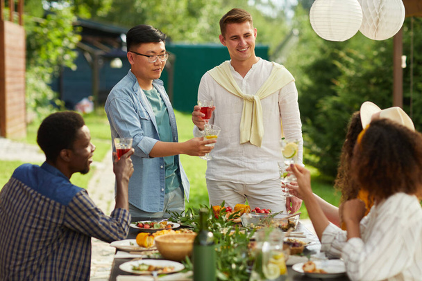 Grupo multiétnico de amigos disfrutando de la cena al aire libre en la fiesta de verano, se centran en dos hombres brindando mientras están de pie junto a la mesa, espacio para copiar - Foto, Imagen